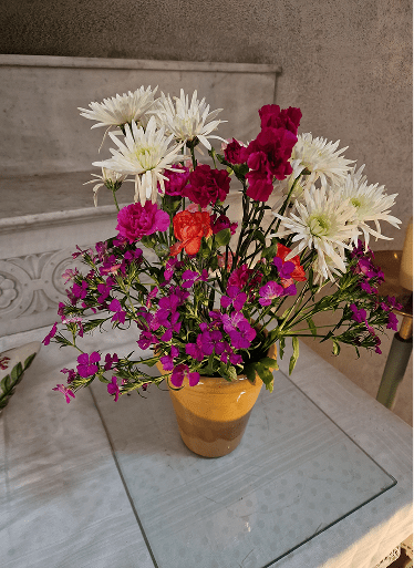 Arrangement floral coloré et touffu présenté dans une poterie jaune, mariant de grands chrysanthèmes blancs échevelés, des œillets rose vif et de petites fleurs violettes sur un plateau de marbre.