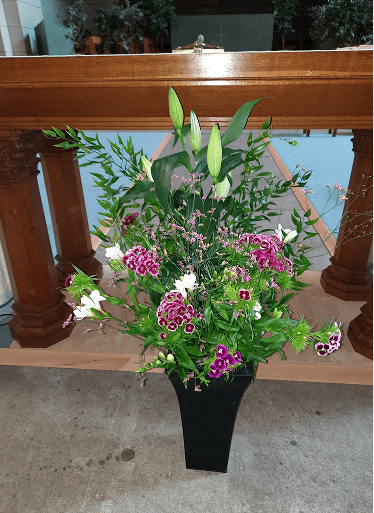 Grand bouquet composé de lys encore en boutons, d'œillets de poète roses et mauves ainsi que de feuillages vert tendre, disposé dans un vase haut noir devant l'autel en bois.