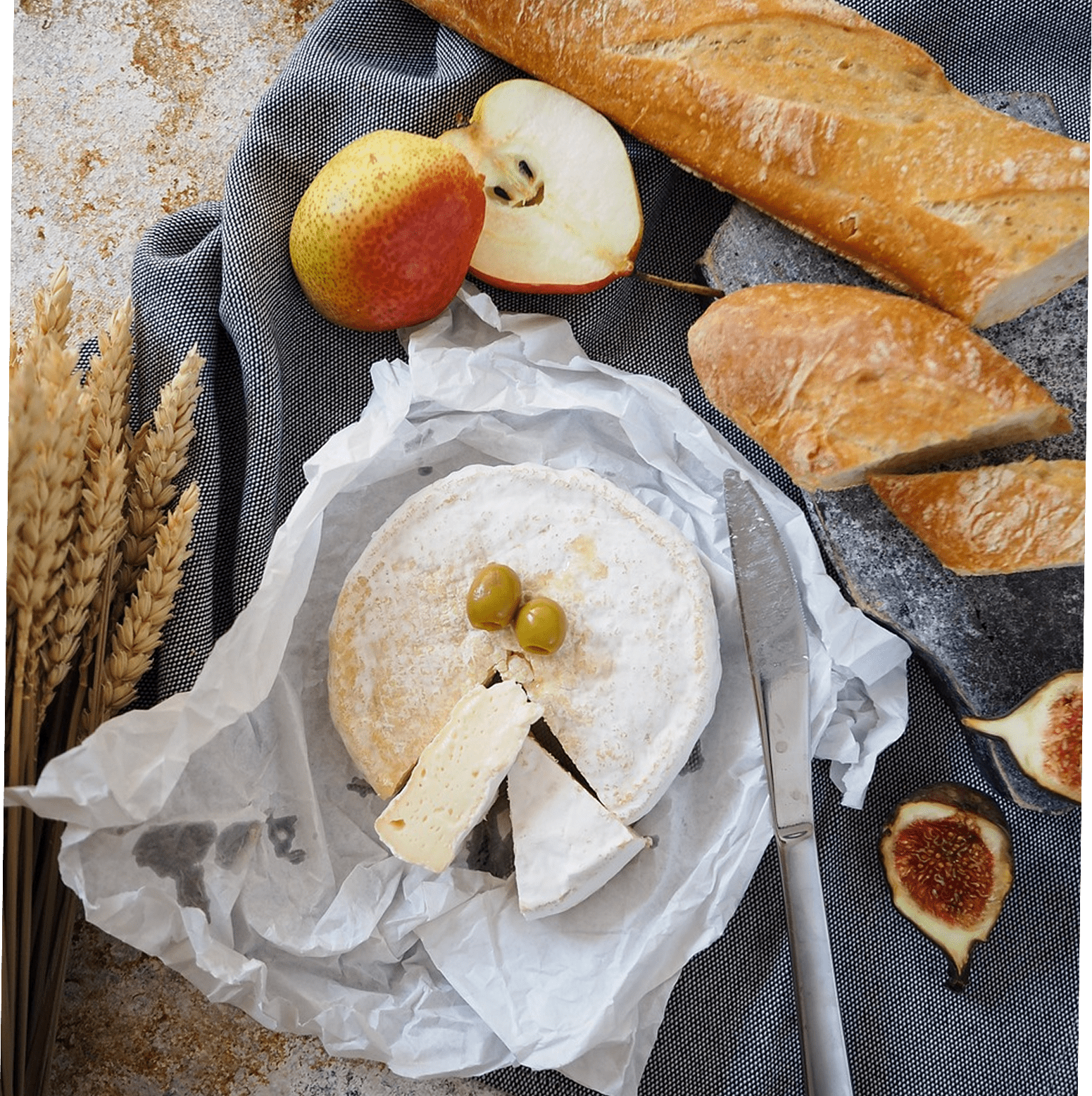 Composition avec un fromage blanc type camembert entamé, du pain baguette, des figues, des poires et des épis de blé.