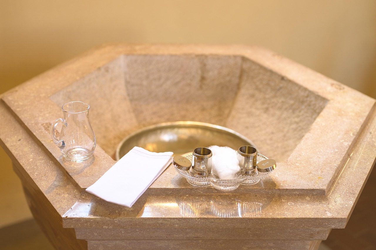Fonts baptismaux et huiles saintes Gros plan sur les fonts baptismaux en pierre claire : sur le rebord sont disposés une aiguière en verre d'eau bénite, un linge blanc (le vêtement blanc) et les flacons du Saint-Chrême et de l'huile des catéchumènes.