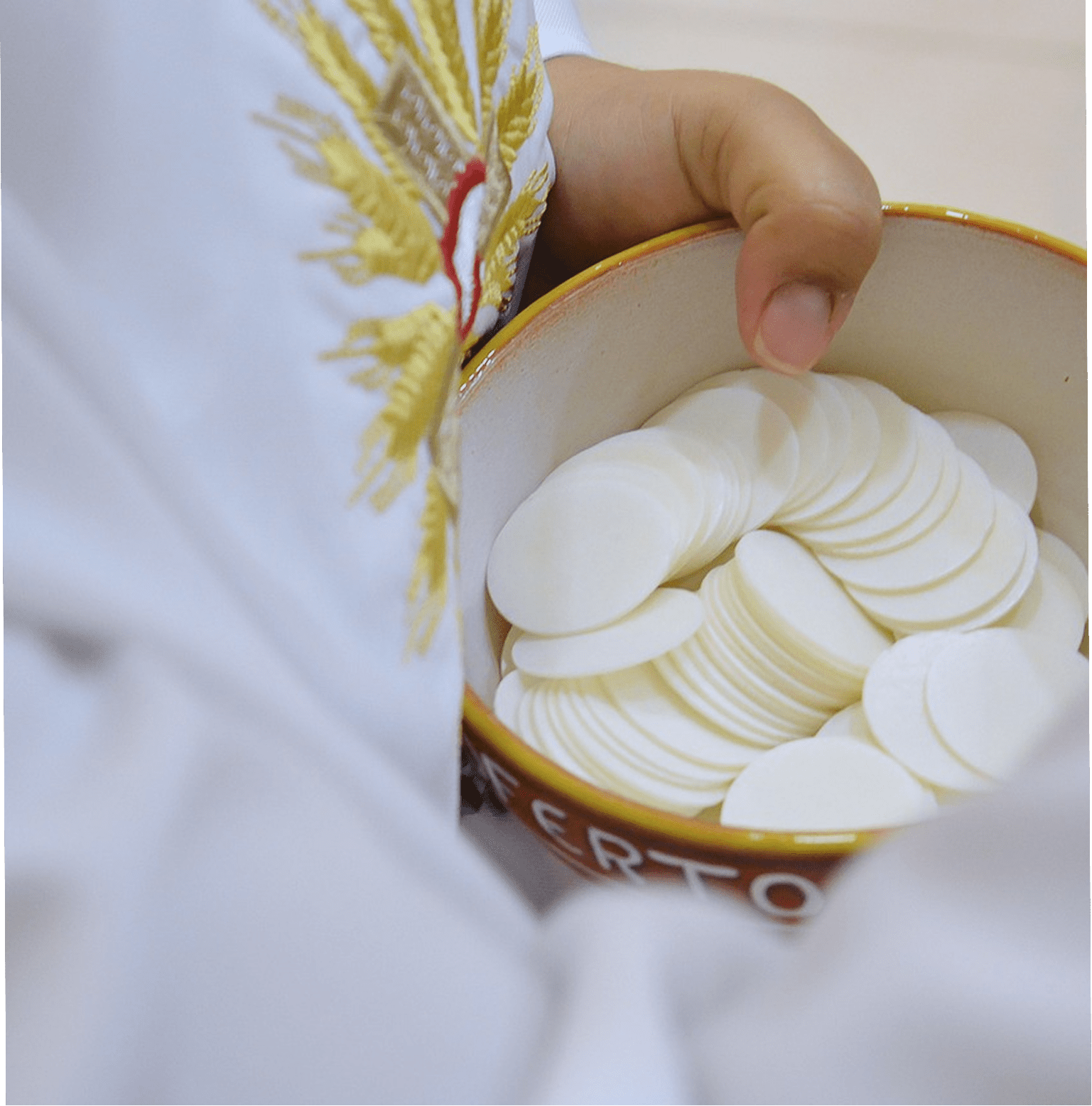 Plan rapproché montrant les mains d'une personne en aube liturgique à broderie dorée, tenant un bol en céramique garni d'hosties blanches.