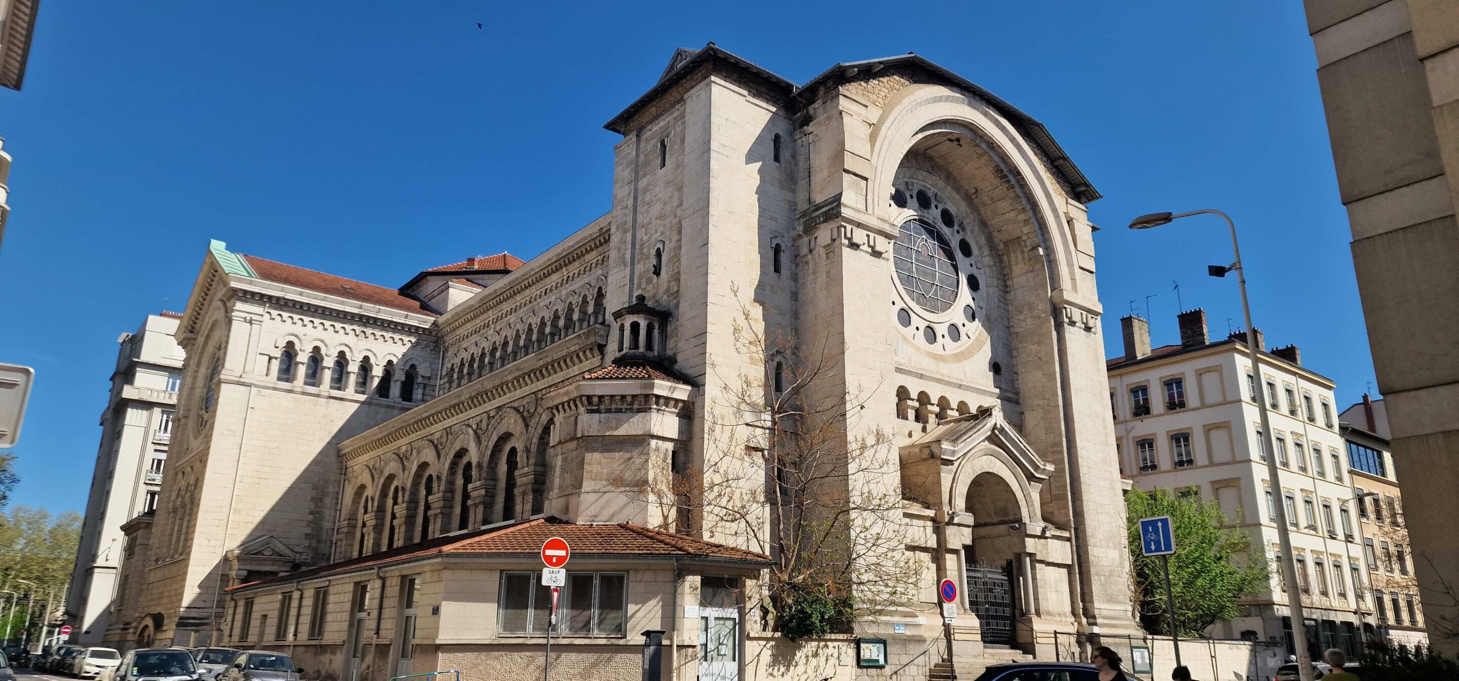 Photographie en contre-plongée de l'architecture extérieure de l'église Saint-Joseph, mettant en valeur la nef en pierre claire, la grande rosace au-dessus du porche d'entrée et le bâtiment annexe sous une vive lumière du jour.