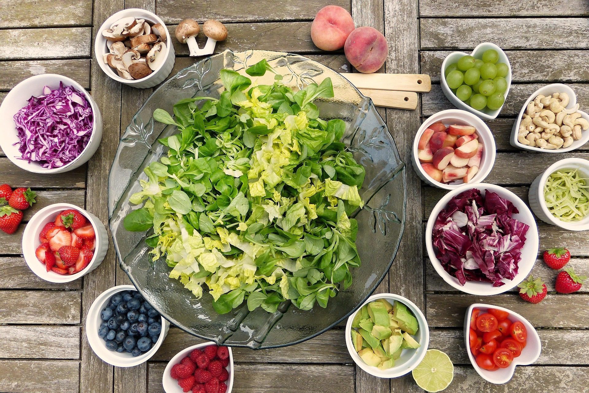 Grand saladier de salade verte entouré de nombreux petits bols d'ingrédients colorés et de fruits sur une table en bois.